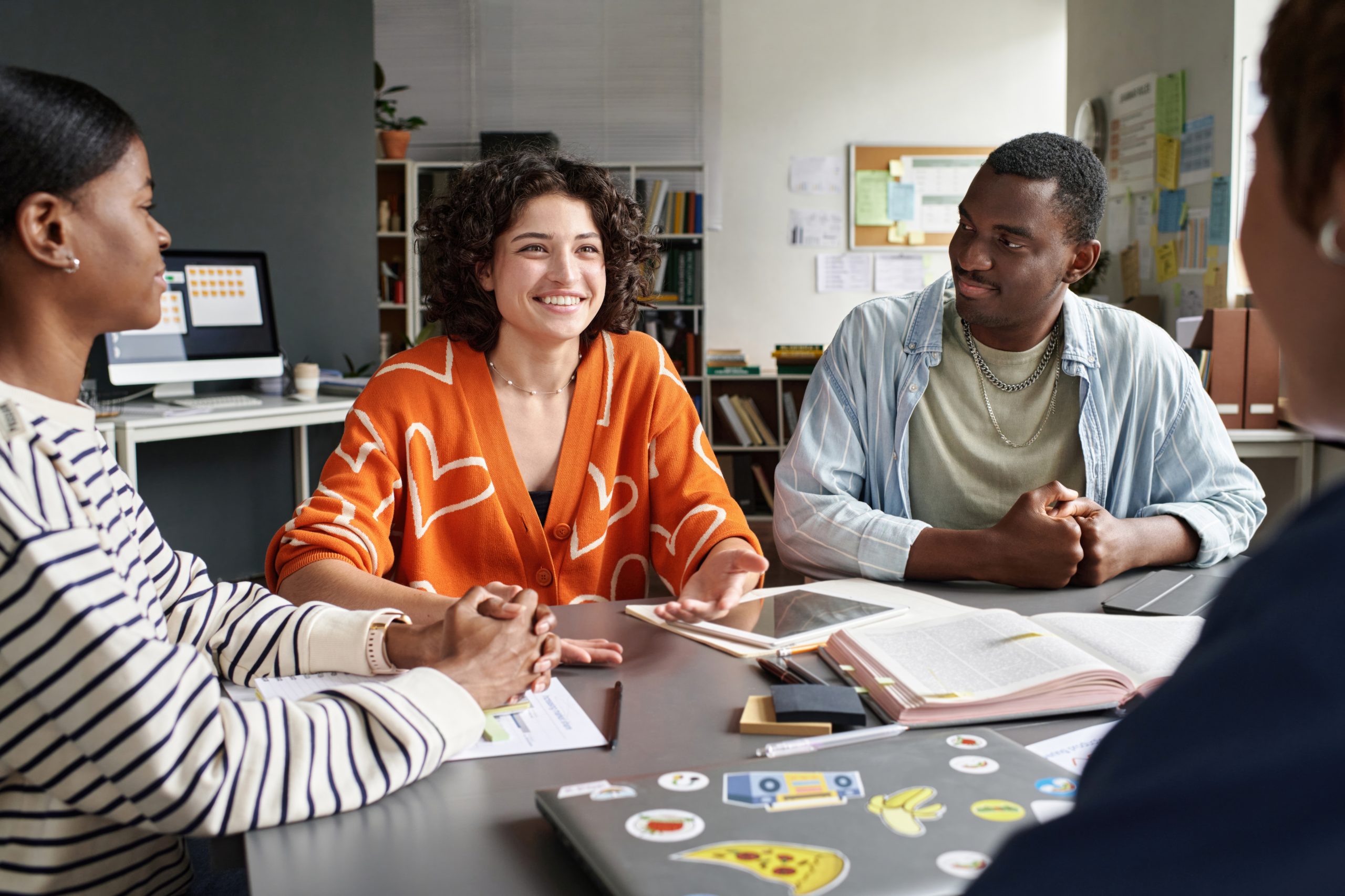 Diverse team discussing project details around office table with laptops and documents visible. Smiling individuals sharing ideas and conducting lively conversation